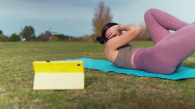 Sporcu kadın mavi yoga minderinde uzanıyor, tablette video izlerken mekik çekiyor.