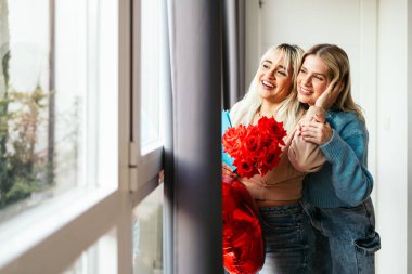 İki sevecen kadın ellerinde kırmızı güller, ellerinde kalp şeklinde bir balon, evde sevgililer gününü kutluyorlar.