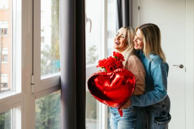 Sevgililer günü için kalp şeklinde bir balonu ve kırmızı gülleri olan bir pencerenin yanında sarılıp gülen mutlu lezbiyen çift.