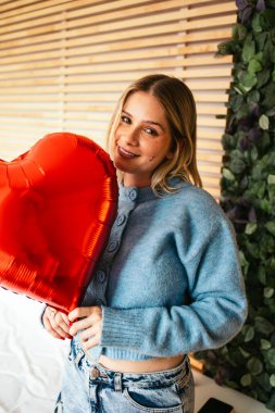 Sevgililer gününü kutlamak için elinde kırmızı kalp şeklinde bir balon tutan gülümseyen genç kadın.