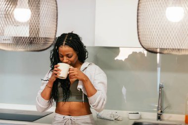 Genç siyah bir kadının şık ve çağdaş mutfağında ılık bir içkinin tadını çıkardığı sakin bir sabah rutini.