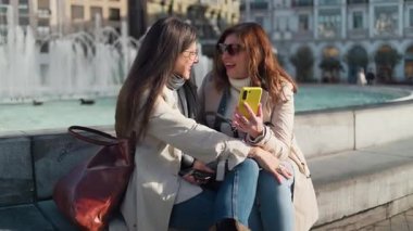 İki olgun bayan arkadaş şehir ortamında cep telefonu kullanarak rahatlatıcı bir günün tadını çıkarıyor.