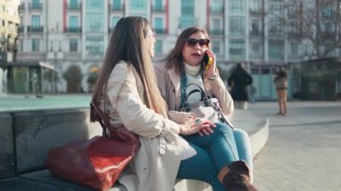 İki neşeli olgun kadın sohbet ediyor, biri cep telefonuyla konuşuyor, şehir meydanında.