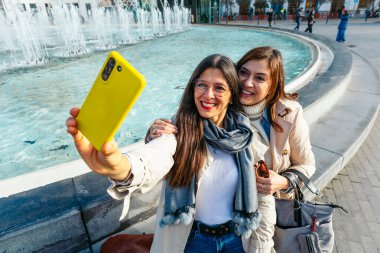 İki neşeli kadın şehir ortamındaki bir çeşmenin yanında selfie çekiyor ve birlikte vakit geçiriyorlar.