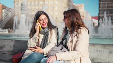 Şehirdeki bir çeşmenin yanındaki bankta oturan iki bayan arkadaş konuşuyorlardı ve akıllı telefon kullanıyorlardı.