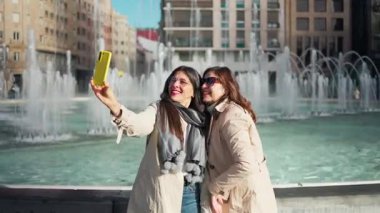 İki neşeli kadın şehir ortamındaki bir çeşmenin yanında selfie çekiyor ve birlikte vakit geçiriyorlar.