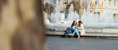 İki bayan arkadaş cep telefonu kullanıyor ve alışveriş çılgınlığından sonra şehir çeşmesinde dinleniyorlar.