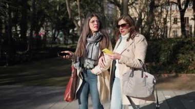 İki bayan arkadaş güneşli bir günün tadını çıkarıyorlar, güzel bir parkta yürüyüp konuşuyorlar.