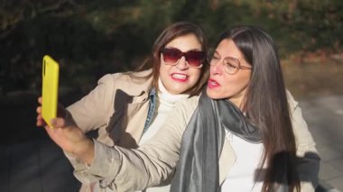 İki olgun kadın, parkta bankta otururken akıllı telefonuyla selfie çekiyor.