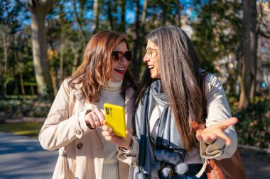 Olgun arkadaşlar kahkahaları paylaşıyor, açık havada akıllı telefon ekranlarına bakıyorlar.