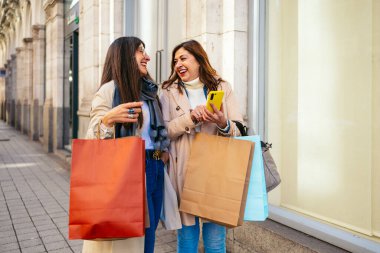 İki olgun kız arkadaş alışveriş yaptıktan sonra sokakta yürüyorlar ve akıllı telefon kullanıyorlar.