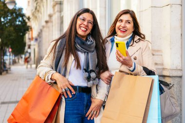 İki mutlu bayan arkadaş alışveriş çılgınlığının tadını çıkarıyor ve şehir ortamında cep telefonu kullanıyorlar.