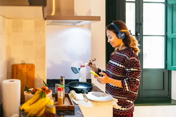 Genç bir kadın kahvaltı hazırlıyor ve kulaklık takıp mutfakta müzik dinlerken akıllı telefon kullanıyor.