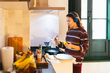 Genç kadın kahvaltı hazırlıyor ve modern mutfakta kulaklık ve pijama giyerken akıllı telefon kullanıyor.