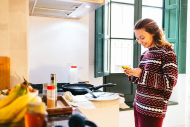 Pijama giyen, akıllı telefon kullanan ve mutfakta kahvaltı hazırlayan genç bir kadın.