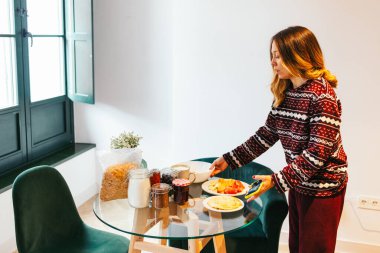 Genç bir kadın kahvaltı için masayı hazırlıyor, pijamalarıyla evde rahat bir sabahın tadını çıkarıyor.