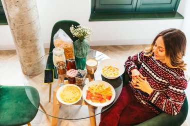 Camdan bir masada oturan genç bir kadın krep, mısır gevreği, meyve ve kahveden oluşan sağlıklı ve çeşitli bir kahvaltı yapıyor.