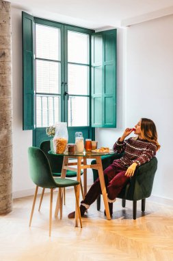 Genç bir kadın pijamalarıyla oturmuş, modern bir dairede yemek masasında sağlıklı bir kahvaltı yapıyor.