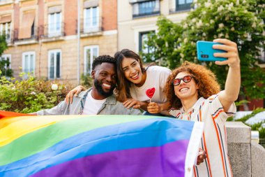 Üç mutlu arkadaş bir gurur etkinliğinde gökkuşağı bayrağı tutarken selfie çekiyorlar, çeşitliliği ve lgbtqia artı hakları kutluyorlar.