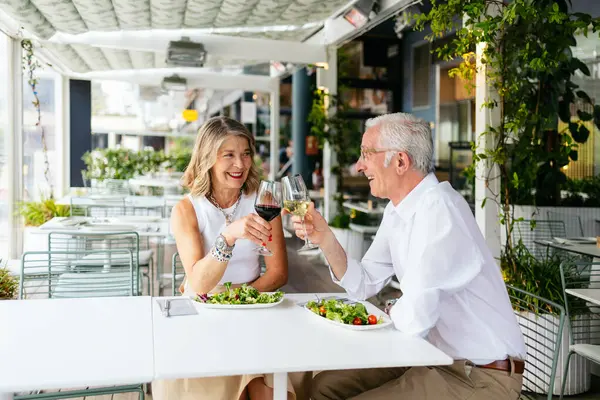 Mutlu son sınıf çifti, açık hava restoranında sağlıklı bir öğle yemeği yerken şarap kadehlerini kaldırıyor..