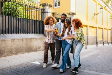 Bir grup çok ırklı arkadaş sokakta yürüyüş yapıyor, bir anı paylaşıyor ve akıllı telefona bakıyor.