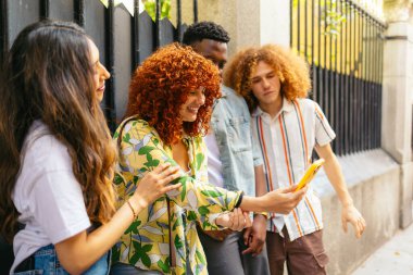 Dört çok ırklı arkadaş açık havada eğleniyor, cep telefonu kullanıyor ve gülümsüyor.