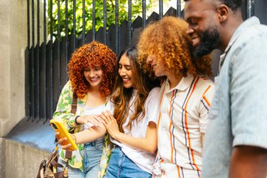 Dört neşeli çoklu etnik arkadaş açık havada akıllı telefon kullanıyor ve birlikte vakit geçiriyorlar.