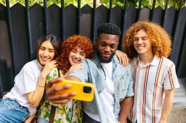 Dört neşeli çoklu etnik arkadaş dışarıda selfie çekiyorlar, arkadaşlığı ve teknolojiyi kucaklıyorlar.