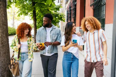 Dört neşeli çoklu etnik öğrenci şehrin kaldırımında yürürken akıllı telefonlar kullanıyor ve boş zamanlarının tadını çıkarıyorlar.