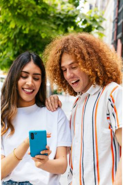 Çok ırklı genç çift, şehir merkezinde bir akıllı telefondan sosyal medya içeriğinin keyfini çıkarıyor.