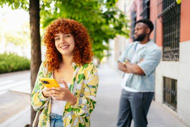 Kızıl saçlı kadın cep telefonu kullanıyor. Sokakta eli kolu çapraz bekleyen bir adam bekliyor.
