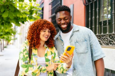 Çok ırklı genç bir adam ve kadın cep telefonuna bakıyor ve şehirde yürürken gülümsüyor..