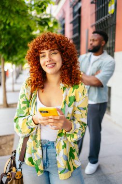 Kıvırcık saçlı, gülümseyen genç bir kadın şehir caddesinde cep telefonu kullanıyor. Arka planda bulanık bir adam var.