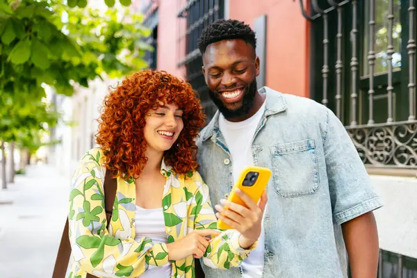 Çok ırklı genç bir adam ve kadın cep telefonuna bakıyor ve şehirde yürürken gülümsüyor..