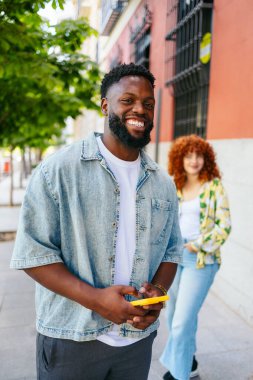 Genç adam gülümsüyor ve cep telefonu kullanıyor. Bir kadın arkadaşıyla sokakta yürürken.