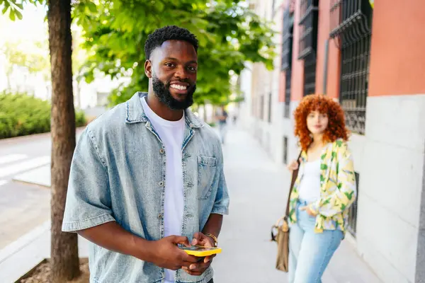 Genç siyahi bir adam, genç bir kadınla şehirde yürürken cep telefonu kullanıyor.