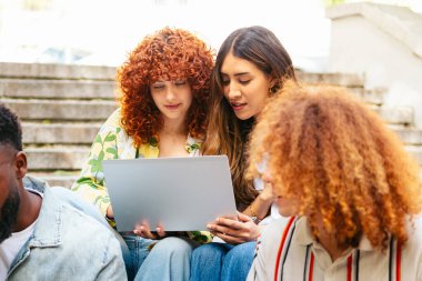 Çeşitli üniversite öğrencileri bir dizüstü bilgisayarda birlikte çalışıyor, dışarıda kampüs basamaklarında ödev yapıyor ve çalışıyorlar.