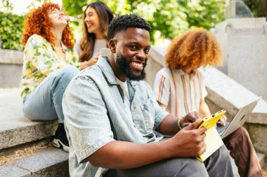 Arkadaşlarıyla dışarıda çalışırken tablet ve laptop kullanan mutlu bir üniversite öğrencisi.