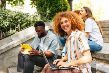 Basamaklarda dizüstü bilgisayar ve dijital tablet kullanarak oturan bir grup öğrenci üniversite kampüsünde açık havada ders çalışmanın keyfini çıkarıyorlar.