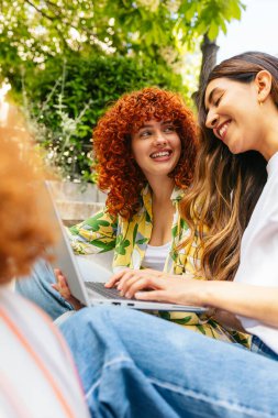 İki kız öğrenci gülümsüyor ve bir dizüstü bilgisayarla kampüste oturuyorlar.