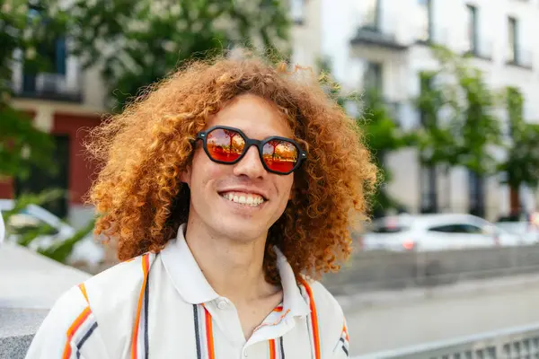 Canlı kırmızı kıvırcık saçlı ve güneş gözlüklü neşeli genç bir adamın portresi, şehirde güneşli bir günün tadını çıkarıyor.