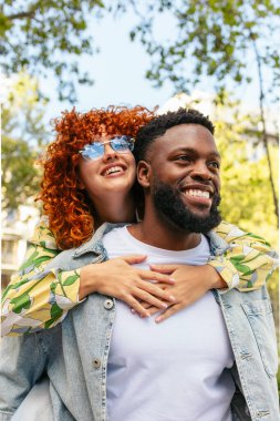Genç kızıl saçlı kadın erkek arkadaşına arkadan sarılıyor ve gülümsüyor, parkta birlikte eğleniyor.