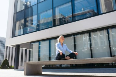 İş kadını yoga ve meditasyon yapıyor ofis binasının önündeki bankta, zihinsel sağlık ve stres yönetimini destekliyor.