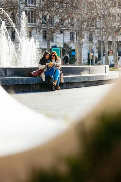 İki bayan arkadaş sosyal medyayı kontrol ediyor ve bir çeşmenin yanında otururken gülümsüyor.