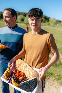 Baba oğul ekmek, peynir ve meyve sepeti taşıyorlar, parkta pikniğin tadını çıkarıyorlar, birliktelik ve sağlıklı yaşam tarzını destekliyorlar.