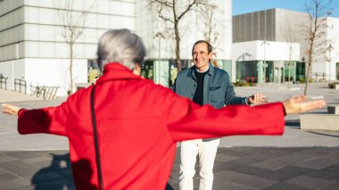 İki olgun arkadaş, şehir meydanında birbirlerini kollarını açarak selamlıyorlar.