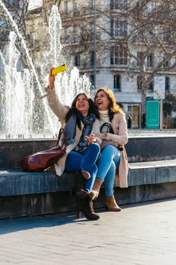 İki neşeli olgun kadın şehirde bir çeşmenin yanında otururken akıllı telefonlarıyla selfie çekiyor.