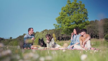 Mutlu bir aile güneşli bir parkta pikniğin tadını çıkarıyor, sevgili köpekleriyle yemek yiyip gülüyorlar.