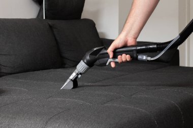 Bir el, gri bir kanepeye ıslak bir elektrikli süpürge takar. Döşemeleri iyice temizler..