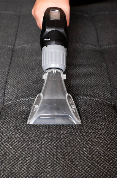 Close-up of a hand holding a transparent wet vacuum cleaner nozzle over a gray sofa, ready for cleaning.